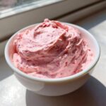A close-up of fluffy pink strawberry cheesecake dip swirled high in a small white bowl, catching bright sunlight.