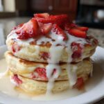 A stack of three fluffy strawberry pancakes topped with fresh sliced strawberries and a generous white cream drizzle.