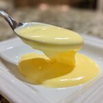 A spoonful of thick, creamy, yellow hollandaise sauce drips onto a white plate.