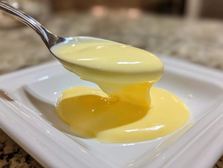A spoonful of thick, creamy, yellow hollandaise sauce drips onto a white plate.