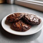 A plate of four rich, dark chocolate brownie cookies featuring shiny, cracked tops.