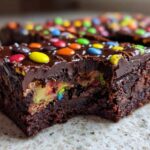 Close-up of a fudgy cosmic brownie square with a bite missing, revealing colorful candy filling and chocolate frosting.