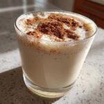 A glass of creamy, chilled coquito topped generously with ground cinnamon spice.