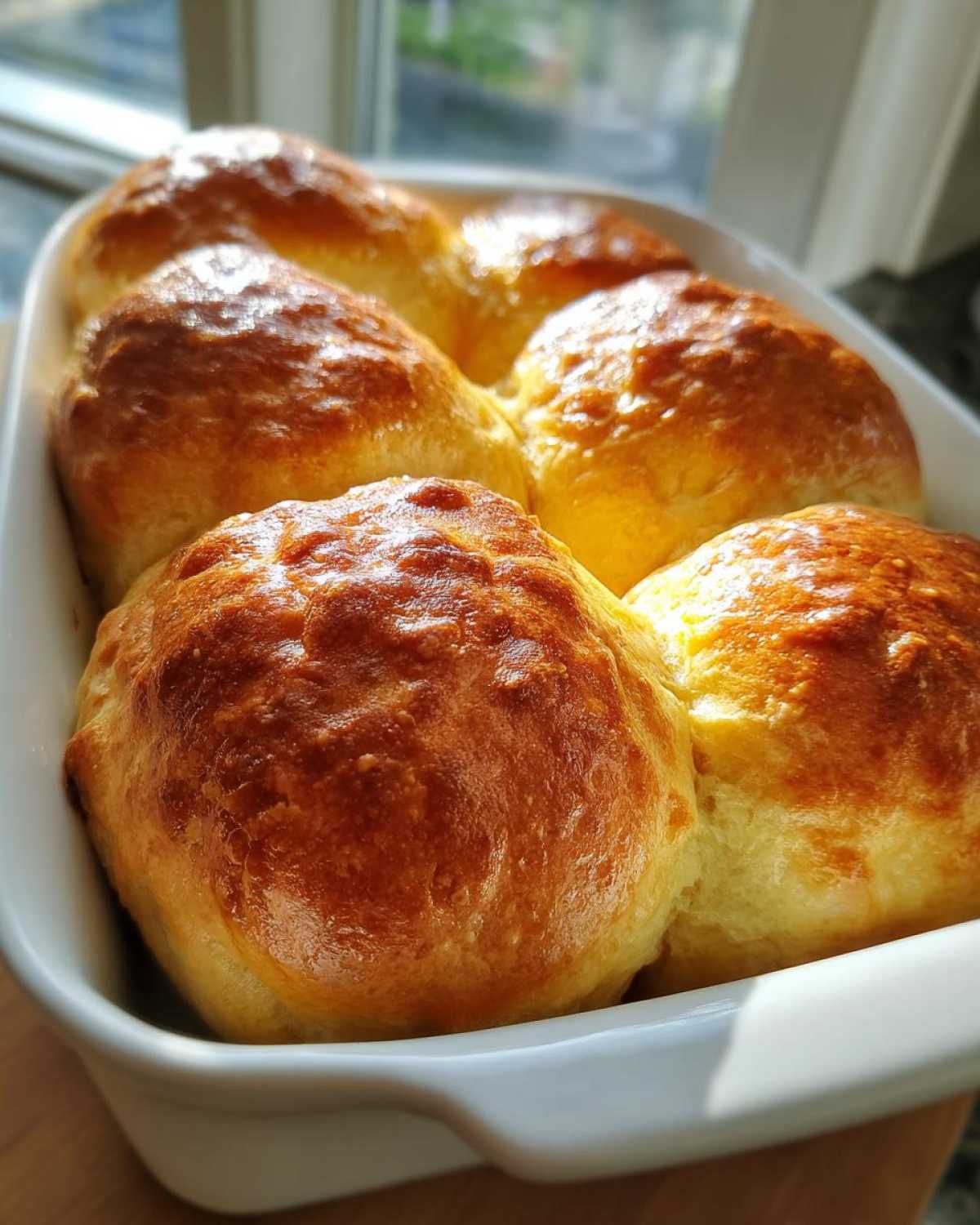 A close-up of six golden brown, shiny sourdough discard rolls baked together in a white ceramic dish.