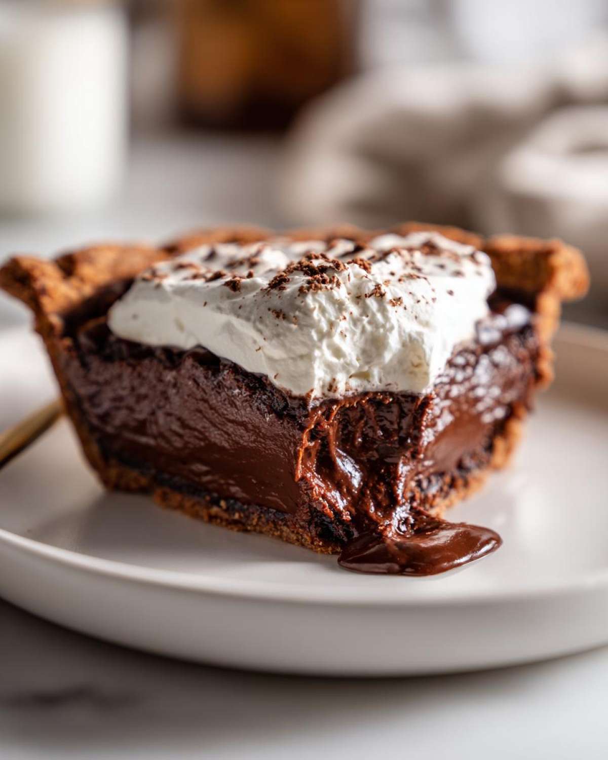 A close-up of a slice of amazing hot fudge pie with a gooey chocolate center oozing onto a white plate, topped with whipped cream.