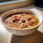 Close-up of a white bowl filled with hearty bean soup featuring ham pieces and mixed beans, lit by sunlight.