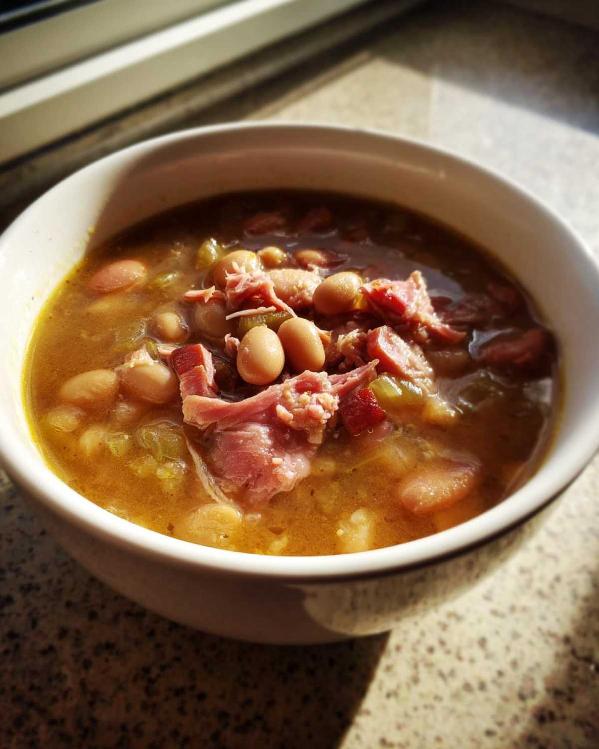 Close-up of a white bowl filled with rich, savory bean soup topped with shredded ham.