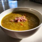 Close-up of a thick, green pea soup in a white bowl, garnished with shredded ham and visible carrot pieces.