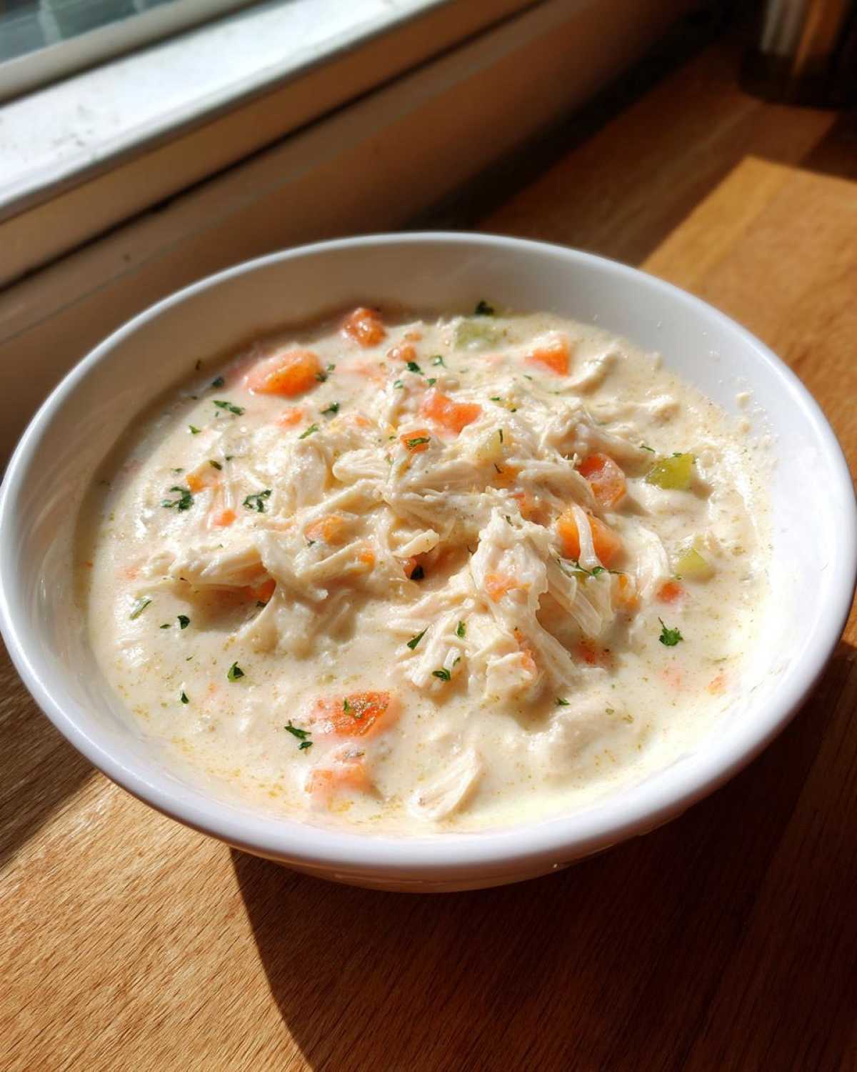 A close-up of a white bowl filled with rich, creamy cream of chicken soup, featuring shredded chicken and diced carrots.
