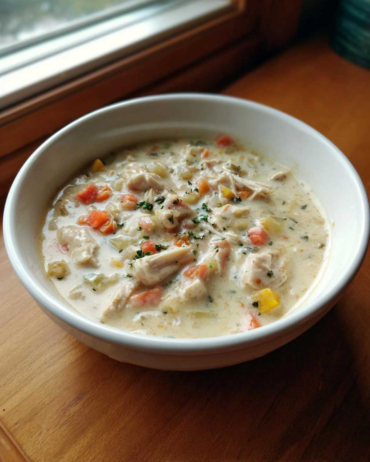 A close-up of a white bowl filled with rich, creamy cream of chicken soup, featuring chunks of chicken and diced carrots.