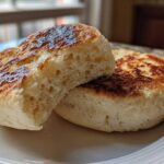 Close-up of a homemade english muffin split in half, showing the airy interior and toasted golden-brown surfaces.