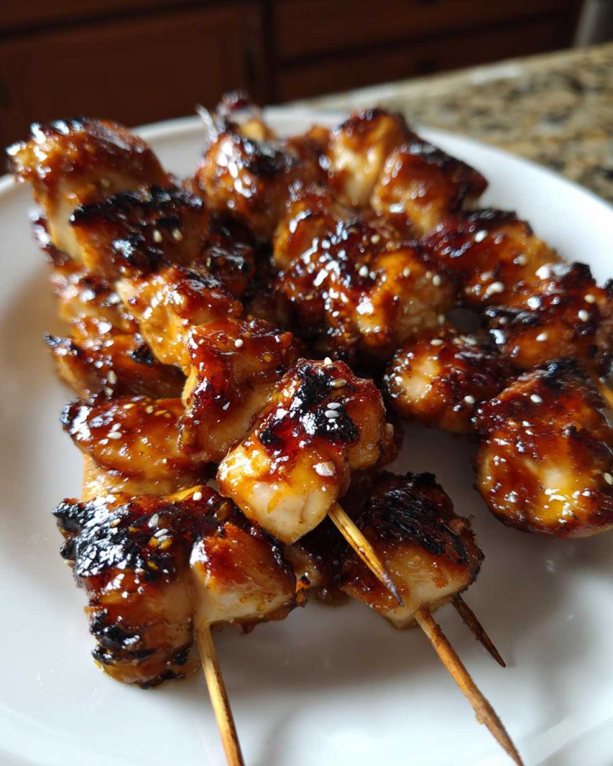 Close-up of several honey garlic chicken skewers sprinkled with sesame seeds on a white plate.