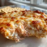 A close-up view of a scoop taken out of a hot, baked reuben dip topped with melted, browned cheese, served with crackers.