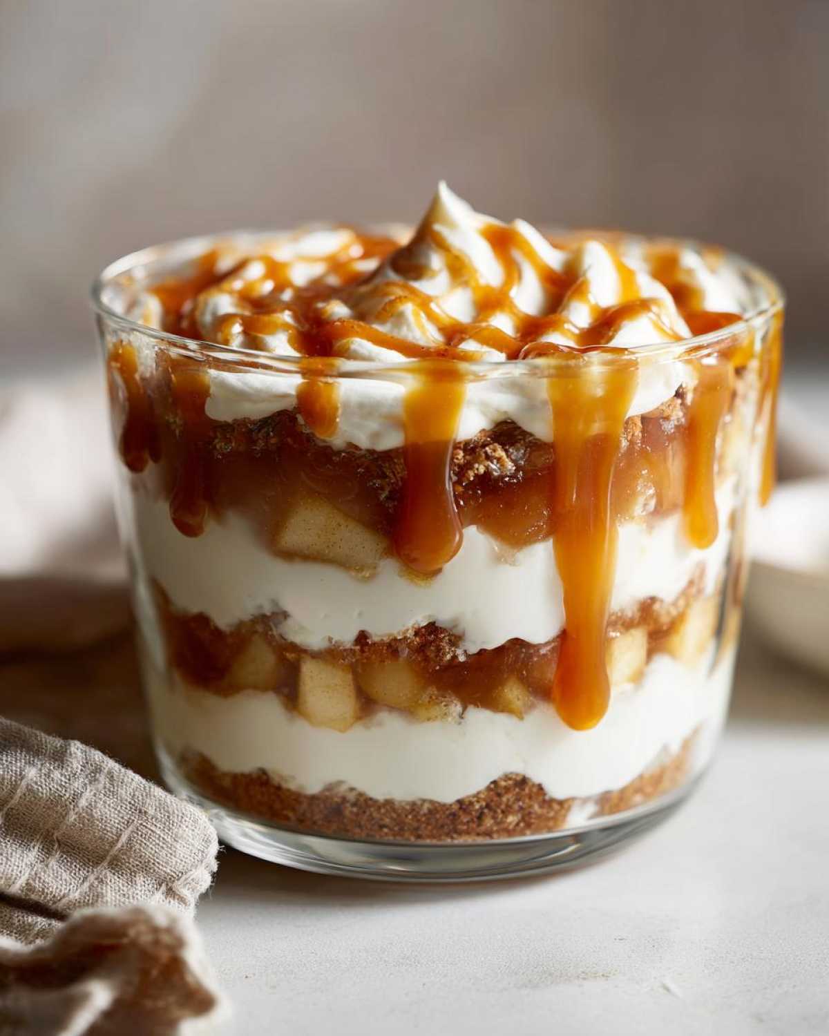Close-up of an individual caramel apple trifle layered with cream, spiced apples, and graham cracker crust, topped with whipped cream and caramel sauce.
