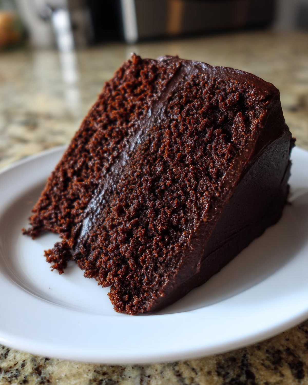 A close-up of a rich, dark slice of moist chocolate cake with thick chocolate frosting on a white plate.