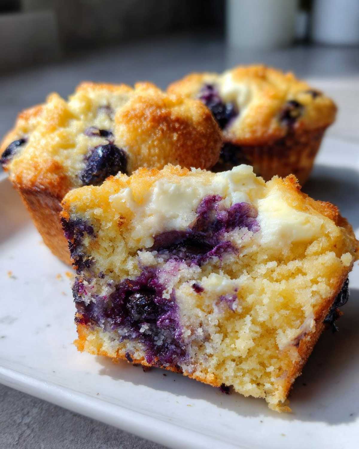 A close-up of a cream cheese muffin cut in half, showing the moist crumb, purple blueberries, and creamy center.