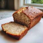 A golden-brown loaf of moist greek yogurt banana bread with one thick slice cut and resting beside it.