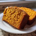 Two thick slices of moist, orange-colored pumpkin bread sitting on a white plate.
