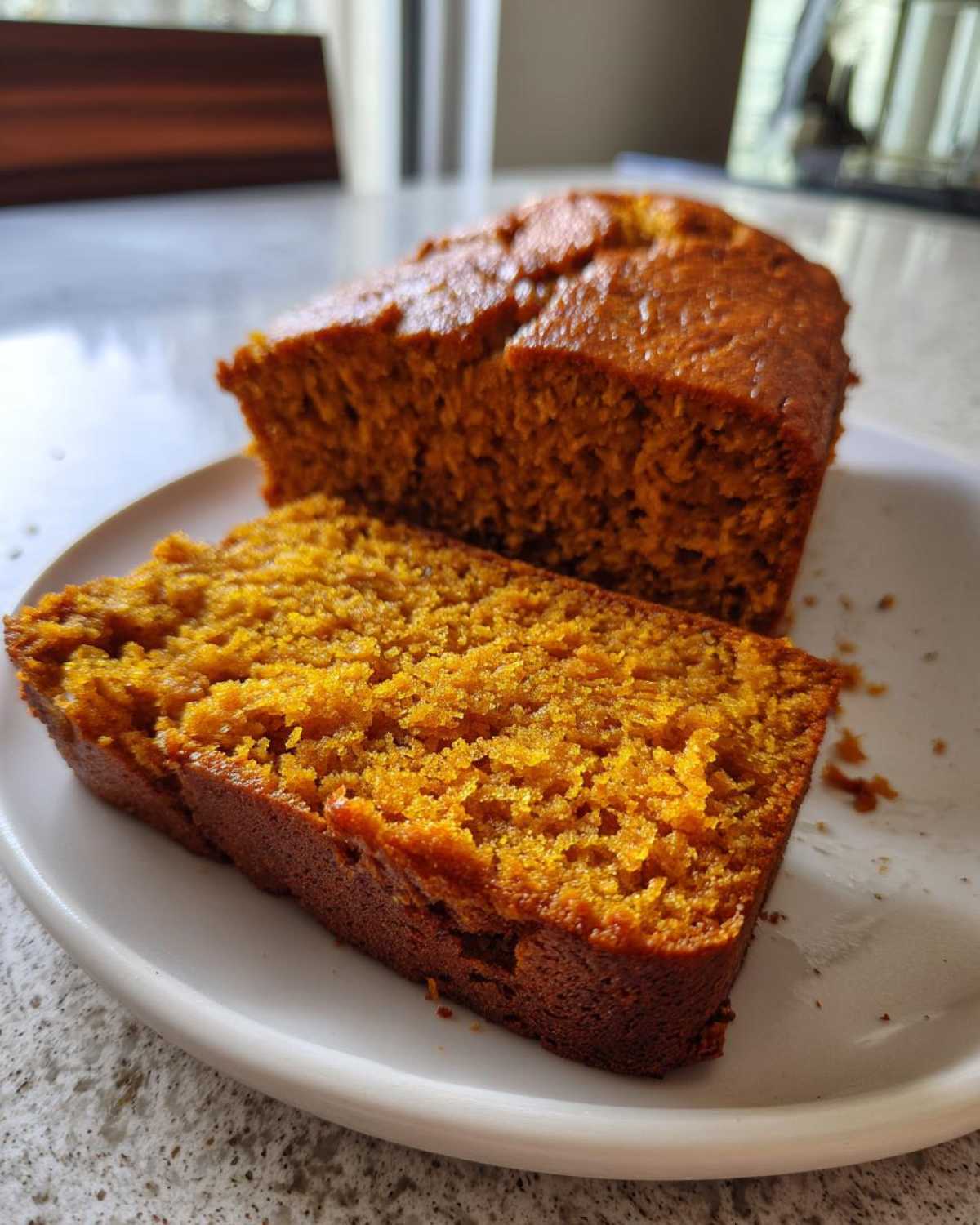 Close-up of a slice of moist pumpkin bread next to the remaining loaf on a white plate.