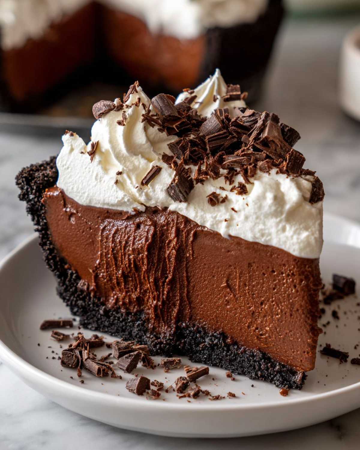 A rich slice of no-bake chocolate mousse pie with a dark crust, topped with whipped cream and chocolate shavings.