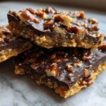 Close-up of stacked squares of rich Pecan Pie Bark showing a crunchy base, dark chocolate layer, and glossy pecans.