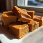 A stack of rich, caramel-colored penuche fudge squares piled on a white serving plate.