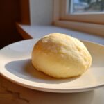 A smooth, pale yellow ball of freshly made fufu resting on a white plate near a sunlit window.