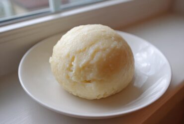 A smooth, round ball of homemade fufu served on a small white plate near a window.