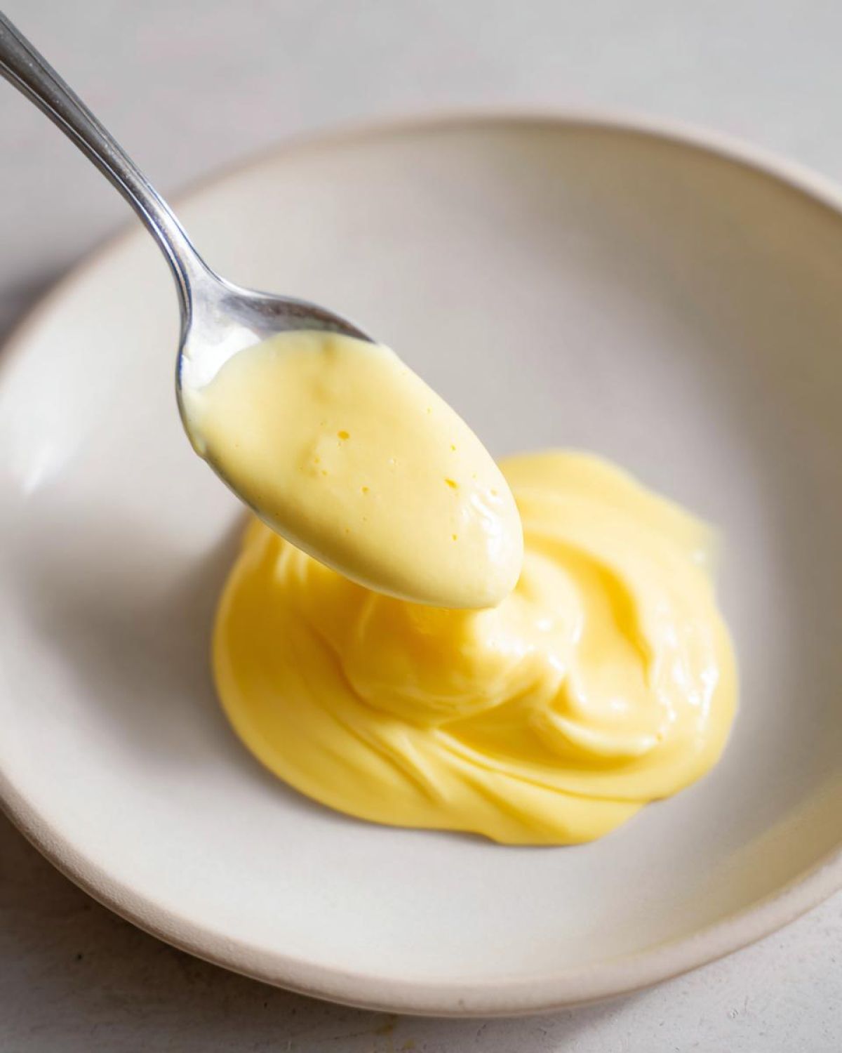 A spoonful of rich, yellow hollandaise sauce being lifted above a dollop of the finished hollandaise sauce on a light plate.