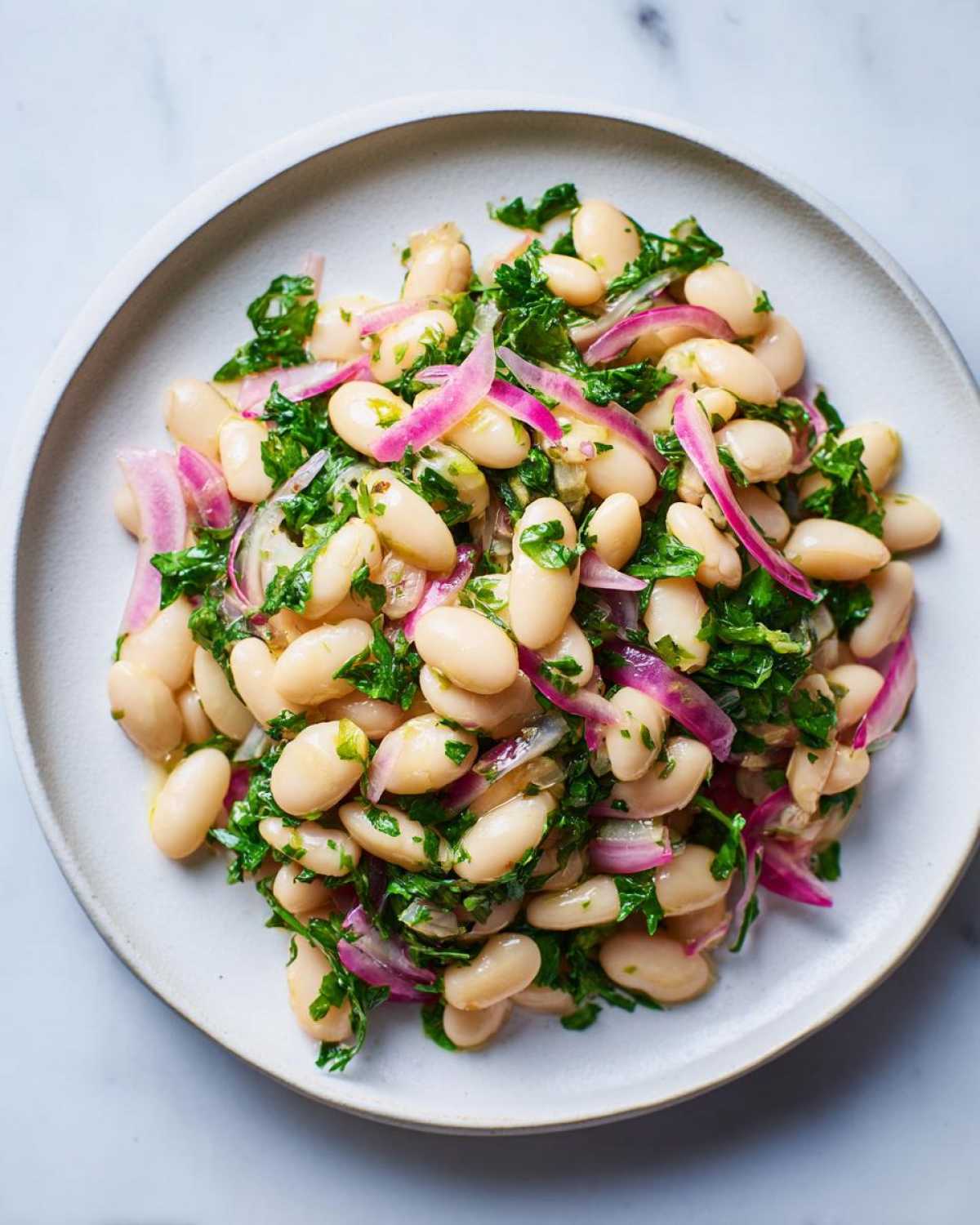 A close-up overhead shot of a vibrant white bean salad featuring cannellini beans, sliced red onion, and fresh parsley.
