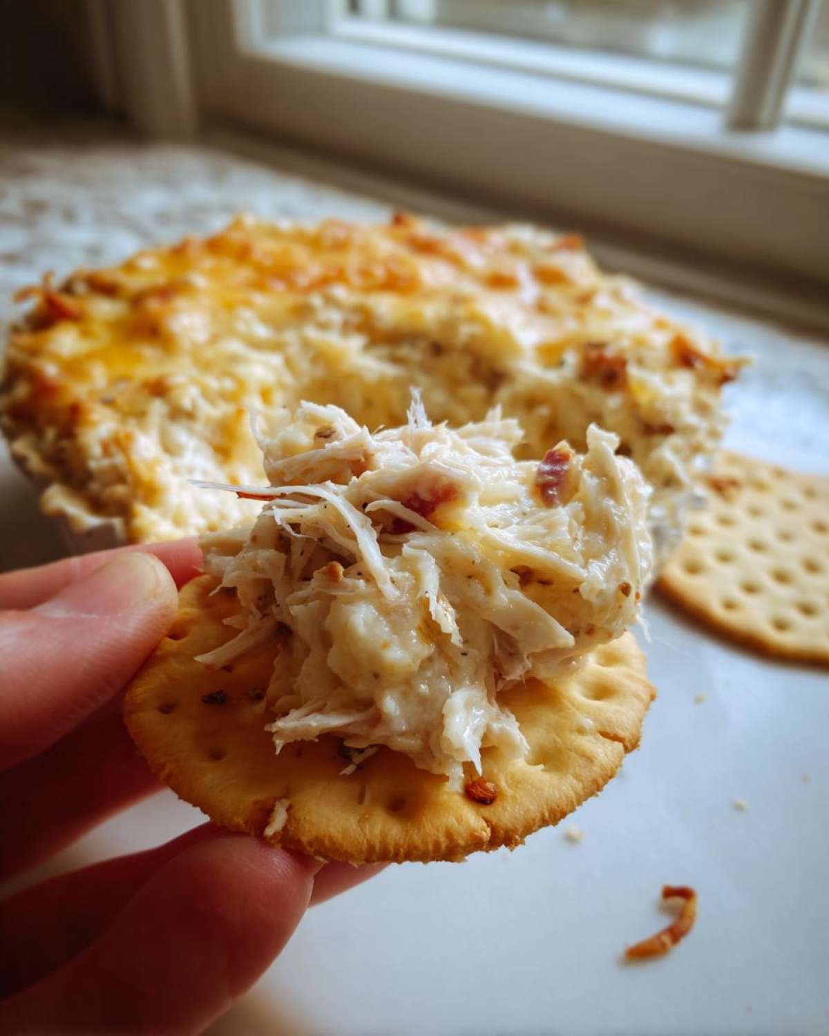 A hand holds a cracker topped with creamy reuben dip, with the baked dip dish visible in the background.