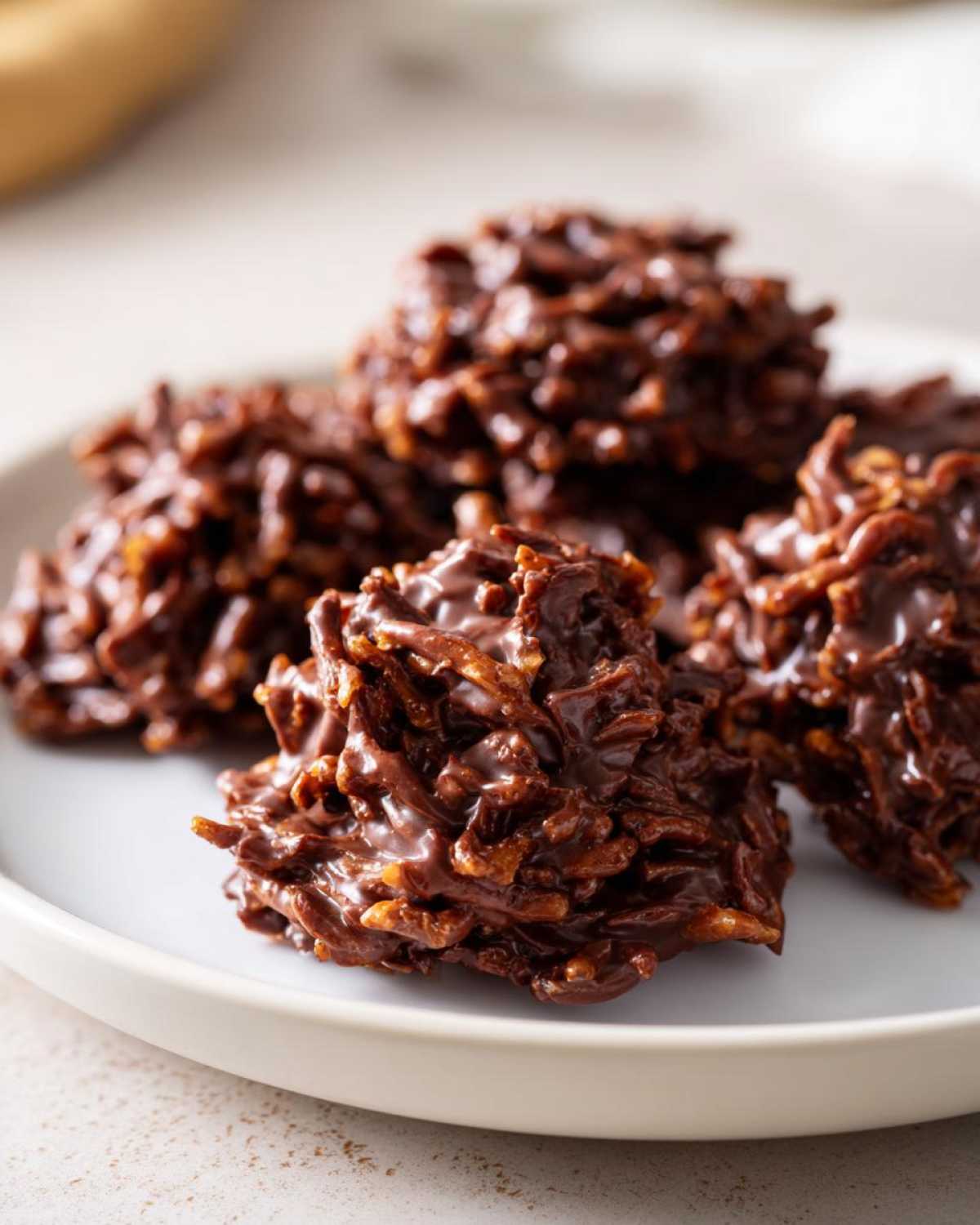 A close-up of several glossy, dark chocolate haystack cookies piled on a light plate.