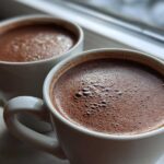 Close-up of two white mugs filled with rich, foamy hot chocolate resting on a windowsill.