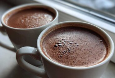 Close-up of two white mugs filled with rich, foamy hot chocolate resting on a windowsill.
