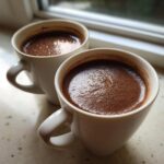 Close-up of two white mugs filled with rich, dark hot chocolate featuring a foamy top layer.