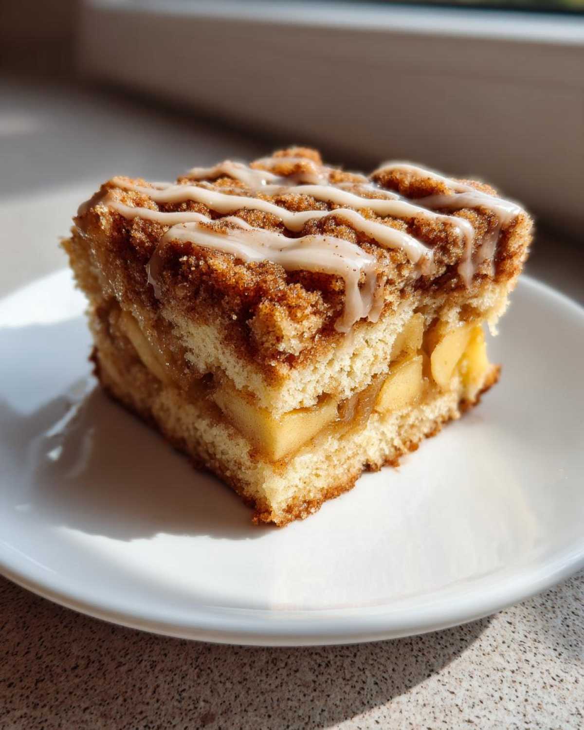 A square slice of moist apple crumb cake topped with cinnamon streusel and a vanilla glaze drizzle.