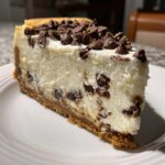 A thick slice of creamy chocolate chip cheesecake with a graham cracker crust and chocolate chips on top.