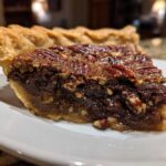 Close-up of a rich slice of chocolate pecan pie showing the flaky crust and gooey filling.