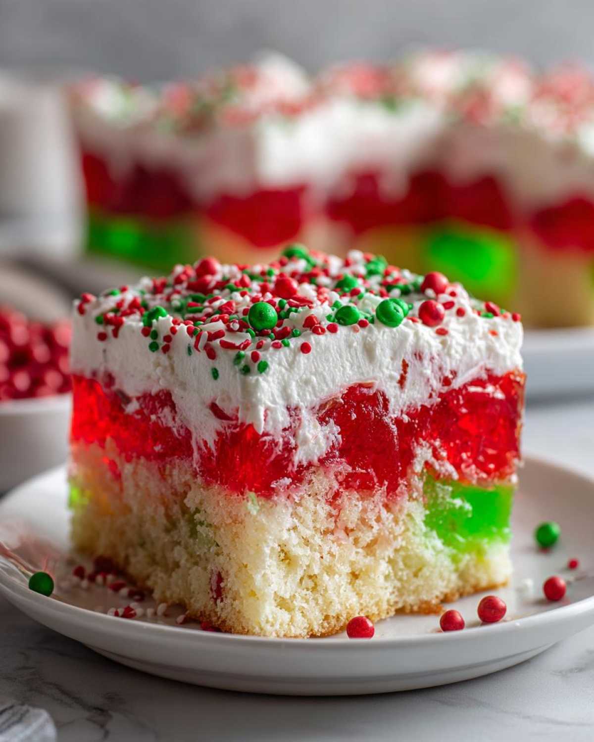 A close-up slice of a vibrant christmas poke cake featuring layers of white cake, red and green jello, whipped topping, and holiday sprinkles.