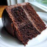 A close-up of a moist slice of homemade chocolate cake covered in dark chocolate frosting on a white plate.