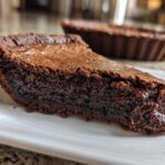 Close-up of a thick slice of dark, gooey fudge pie resting on a white plate, with the rest of the pie in the background.