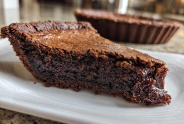 Close-up of a thick slice of dark, gooey fudge pie resting on a white plate, with the rest of the pie in the background.