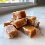 A stack of soft, chewy homemade caramel squares, individually wrapped in plastic wrap, resting on a white marble surface.