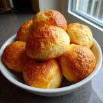 A white bowl filled with golden brown, soft sourdough discard rolls stacked high, sitting near a window.