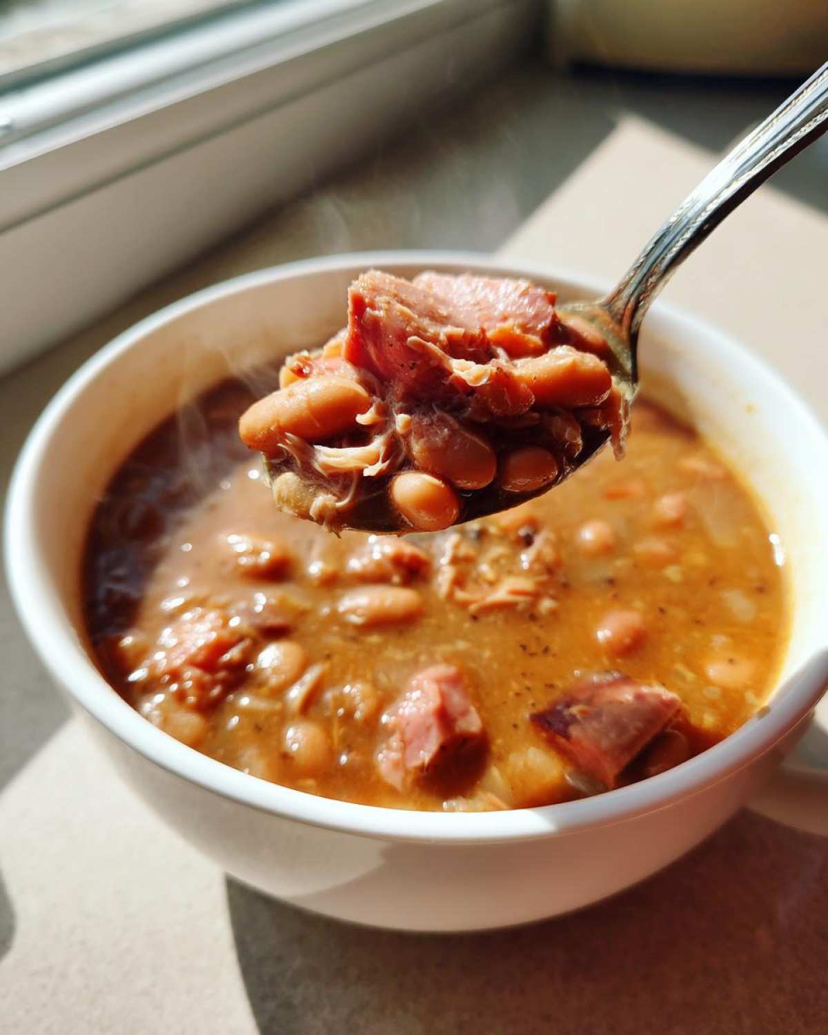 A spoonful of hearty, steaming bean soup with chunks of ham being lifted from a white bowl.
