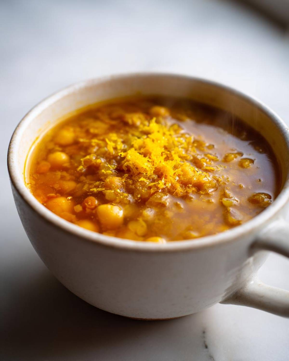 Close-up of a steaming white mug filled with vibrant yellow chickpea soup, topped with bright orange zest.