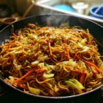 Close-up of delicious, steaming fried noodles mixed with cabbage and carrots cooking in a black skillet.