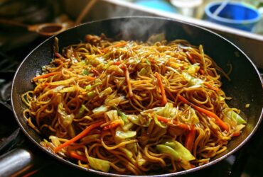 Close-up of delicious, steaming fried noodles mixed with cabbage and carrots cooking in a black skillet.