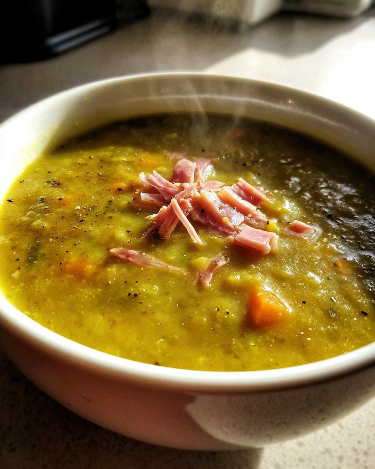 Close-up of a steaming bowl of thick, yellow-green pea soup topped with shredded ham and black pepper.