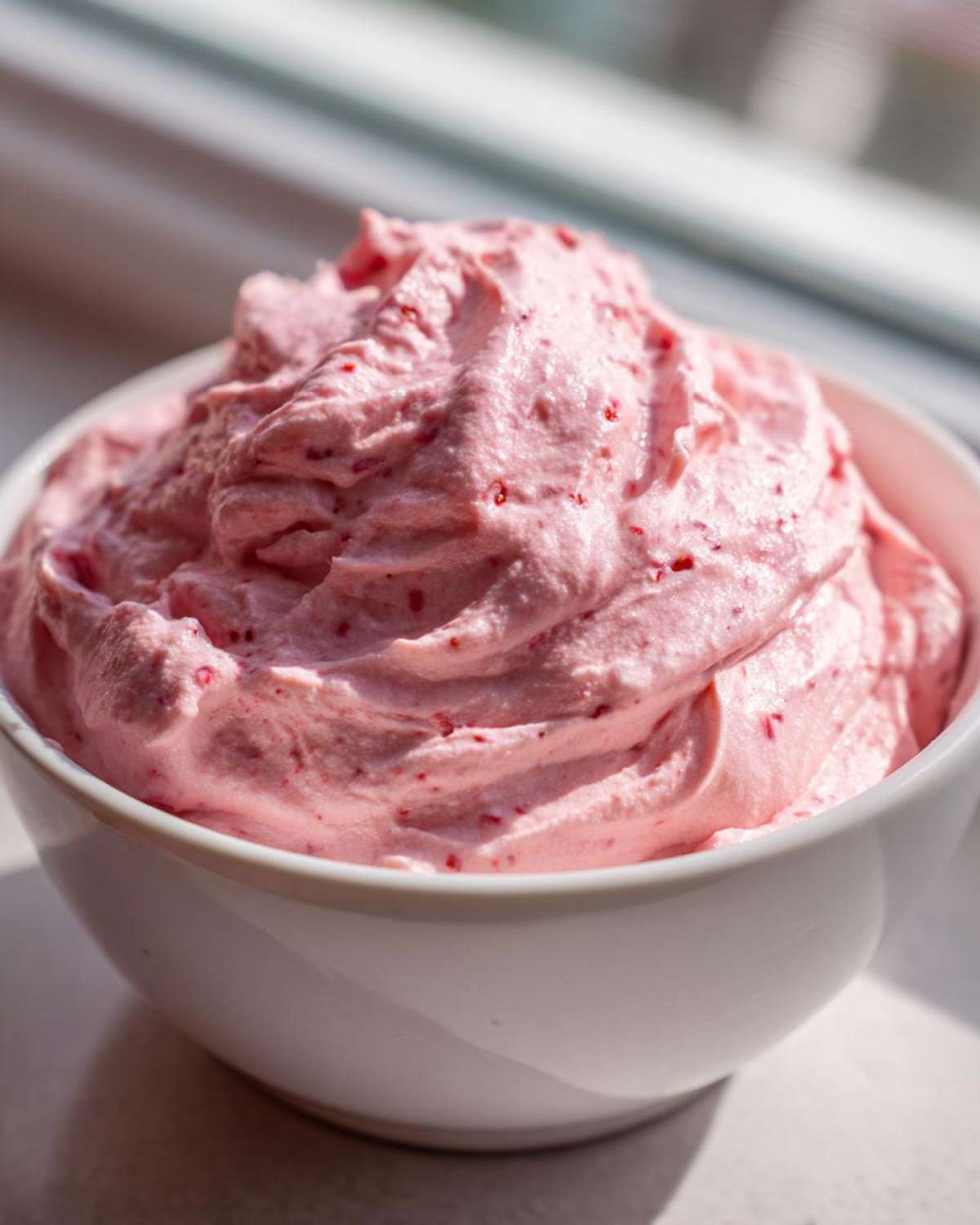 Close-up of fluffy, pink strawberry cheesecake dip swirled high in a white bowl, showing flecks of strawberry.